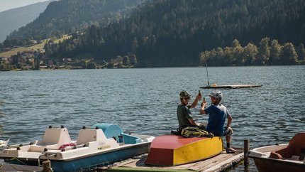 Carinthia Wasserreich Tour © David Karg Two cyclists on dock at lake with mountains in background toasting bottles