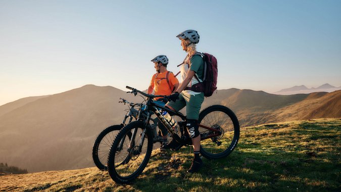 Mountain Bike Holidays © Tobias Köhler Zwei Mountainbiker mit Helmen stehen mit Fahrrädern auf einem Berg bei Sonnenuntergang