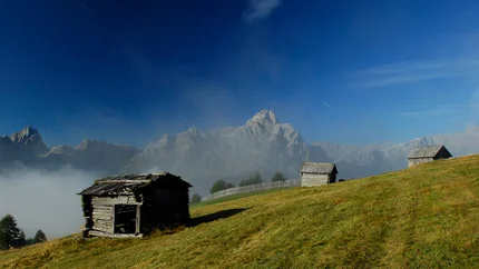 PLÄTZWIESEN TOUR © Harald Wisthaler Alpine huts on green meadow with misty mountains under blue sky