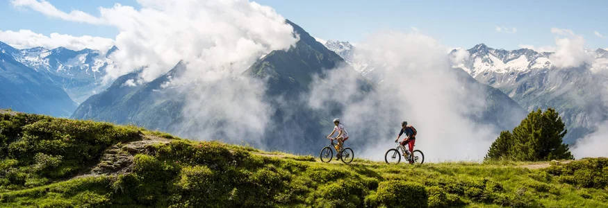 Zillertal © TVB Mayrhofen - Michael Werlberger Two cyclists on a mountain trail with clouds and snow-covered peaks in the background