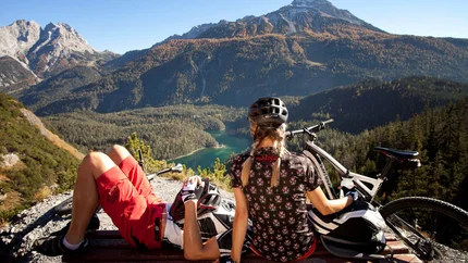 FROM SCHÖNRUH AROUND THE DANIEL © Sporthotel Schönruh Two mountain bikers resting on bench overlooking mountains and lake