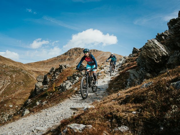 Quality Promise © Desire Lines - Tobias Dörr Two mountain bikers ride on a rocky mountain trail under a blue sky