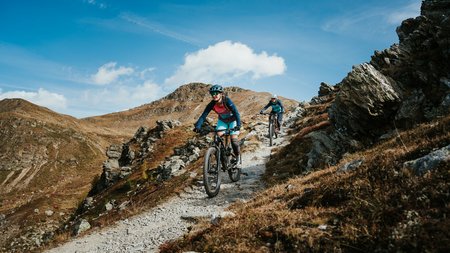 Quality Promise © Desire Lines - Tobias Dörr Two mountain bikers ride on a rocky mountain trail under a blue sky