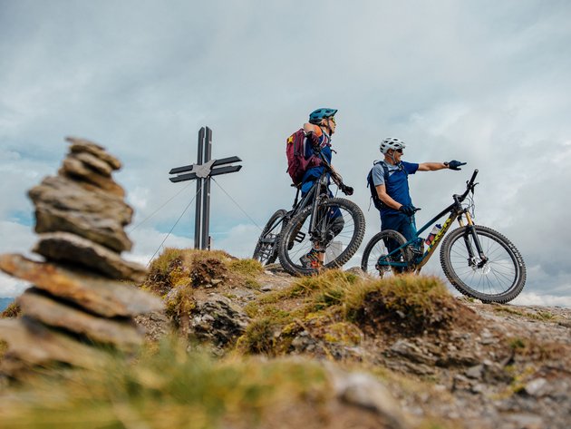 Quality Promise © Felix Saller Two cyclists with helmets standing on a mountain peak near a cross
