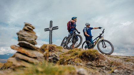 Quality Promise © Felix Saller Two cyclists with helmets standing on a mountain peak near a cross