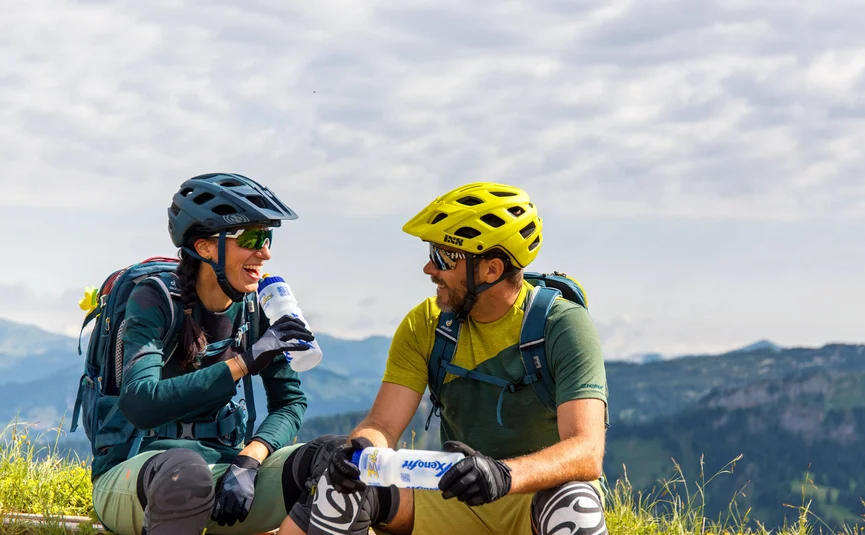 Diedamskopftrail © David Karg Two cyclists taking a break laughing on a mountain meadow with water bottles
