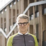 Mountain Bike Holidays © Hannes Niederkofler Älterer Mann in Fahrradbekleidung hält Helm vor einem modernen Gebäude