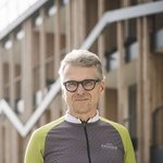 Mountain Bike Holidays © Hannes Niederkofler Älterer Mann in Fahrradbekleidung hält Helm vor einem modernen Gebäude