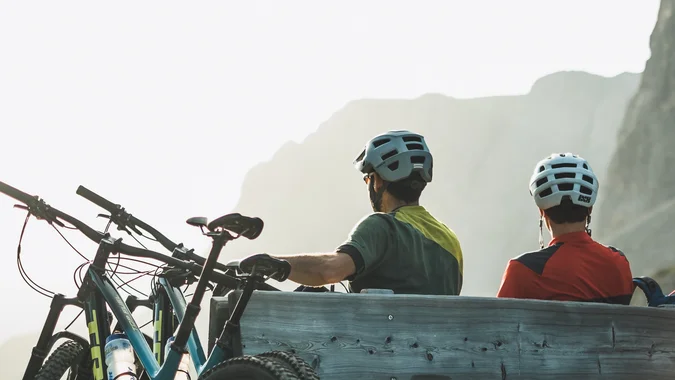 Mountain Bike Holidays © Norman Bielig Zwei Radfahrer mit Helmen sitzen auf einer Holzbalkenbank vor Berglandschaft