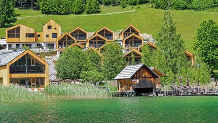 See-Aktivhotel Regitnig - Hotel & Chalets © Regitnig GmbH Holzferienhäuser am See mit grünen Bäumen und Hügeln im Hintergrund