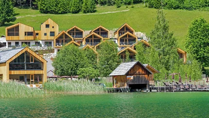 See-Aktivhotel Regitnig - Hotel & Chalets © Regitnig GmbH Holzferienhäuser am See mit grünen Bäumen und Hügeln im Hintergrund