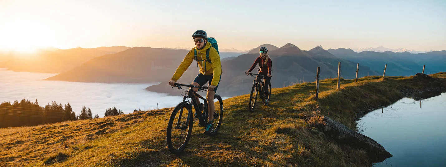 HAHNENKAMM © TVB Kitzbüheler Alpen Brixental - Mathäus Gartner Two mountain bikers cycling at sunrise on a hill with mountain view