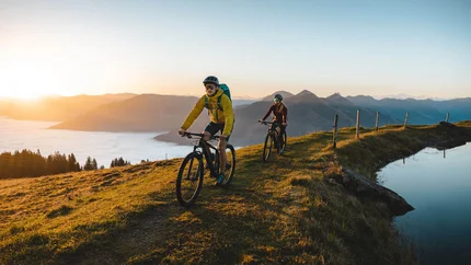 HAHNENKAMM © TVB Kitzbüheler Alpen Brixental - Mathäus Gartner Two mountain bikers cycling at sunrise on a hill with mountain view