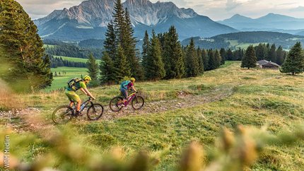 Dolomites - Val d'Ega © WOM Medien - Andreas Meyer Two mountain bikers cycling on a trail in the mountains with fir trees and a cabin