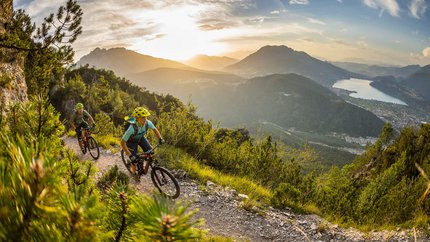 GIRO DELLA MARZOLA © WOM Medien - Andreas Meyer Two mountain bikers riding on a mountain trail at sunset with lake view