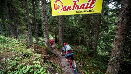 Rauher Kopf - Suntrail © TVB Kitzbühler Alpen - Erwin Haiden Two mountain bikers riding on a forest trail under an OurTrail banner