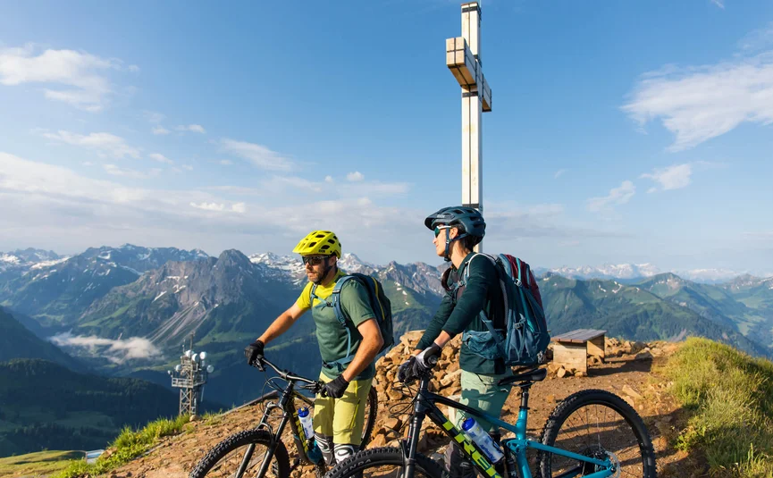 Diedamskopftrail © David Karg Two mountain bikers with helmets at summit cross in alpine mountains