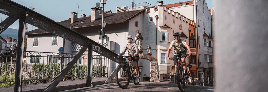 Bressanone © Brixen Tourismus - Hannes Engl Two cyclists riding on a bridge in a historic town on a sunny day