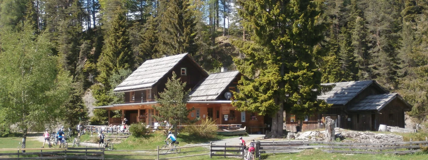 Naggler Alm Tour © Regitnig GmbH Wooden cabins in forest with cyclists on grassy meadow