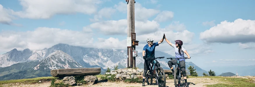 Flachau © Tobias Köhler Two mountain bikers high-five at a summit cross in the mountains