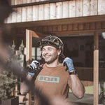 Mountain Bike Holidays © Manuel Kottersteger Mann mit Fahrradhelm und Handschuhen lacht vor einem Hotel mit Holzeingang