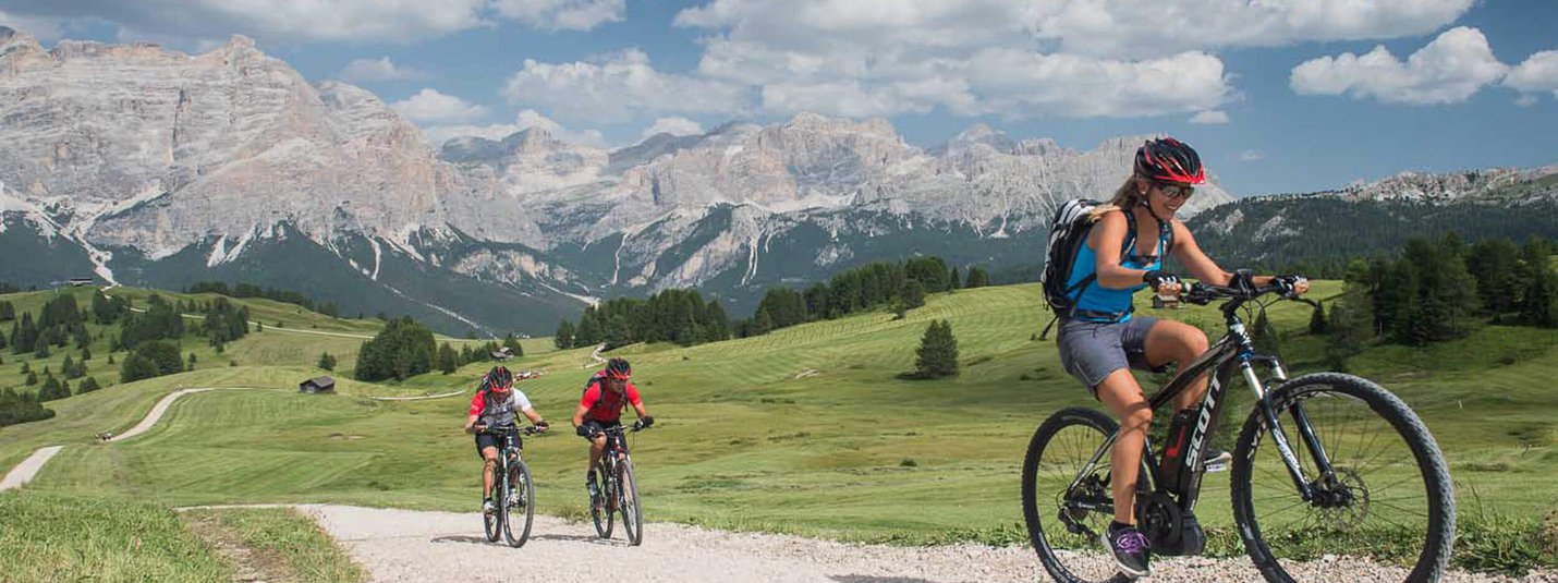 FANES-SENNES ROUND © Excelsior Dolomites Life Resort Three cyclists on gravel path with mountain backdrop on a sunny day
