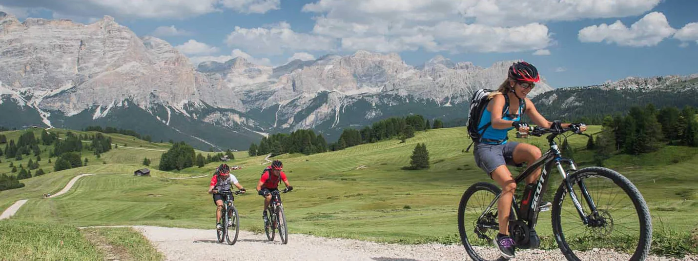 FANES-SENNES ROUND © Excelsior Dolomites Life Resort Three cyclists on gravel path with mountain backdrop on a sunny day