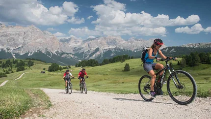 FANES-SENNES ROUND © Excelsior Dolomites Life Resort Three cyclists on gravel path with mountain backdrop on a sunny day