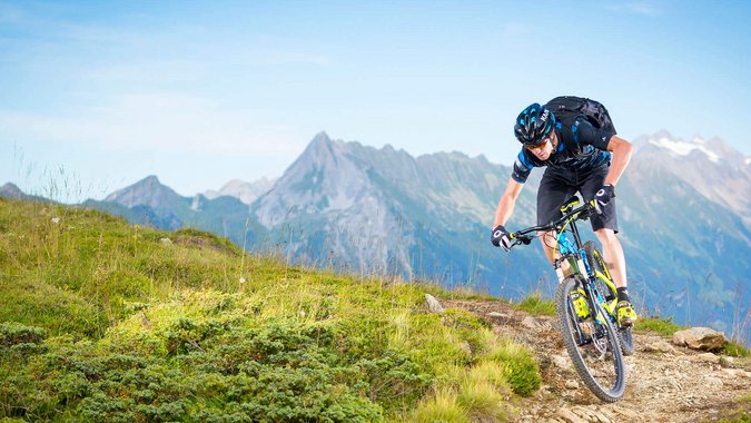 Mountain Bike Holidays © Daniel Geiger Mountainbiker fährt auf Gebirgspfad mit Alpen im Hintergrund