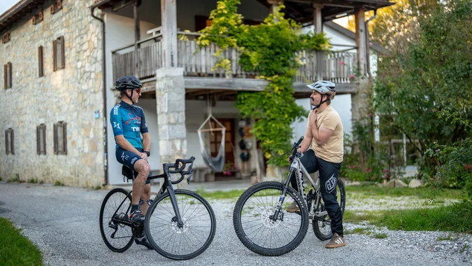 Mountain Bike Holidays © Ziga Koren Zwei Männer mit Fahrrädern stehen auf einem Weg vor einem Haus und unterhalten sich
