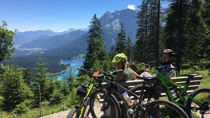 FROM SCHÖNRUH AROUND THE THANELLER © Sporthotel Schönruh Two cyclists sitting on bench overlooking mountain and lake in Alps