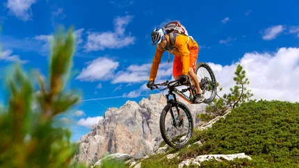 SELLARONDA (CLOCKWISE) © Davide dal Mas Mountain biker in orange gear jumps on rocky alpine trail under sunny sky