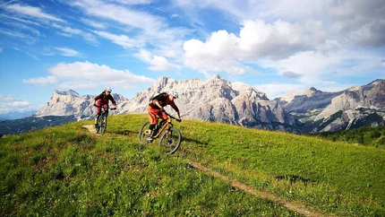 FANES CIRCUIT © Markus Rubatscher Two mountain bikers riding on a trail through green meadow with rocky mountains