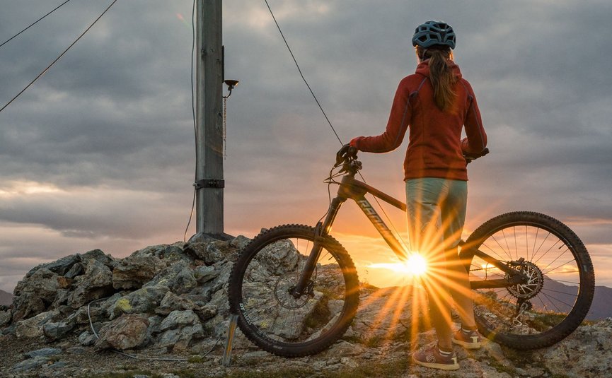 Mountain bike holidays in the 3 Zinnen Dolomites region © Elias Ochner - Stefan Schwabl Person with bike standing next to summit cross at sunset