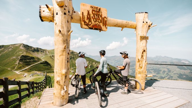 Unterwegs am Schattberg © saalbach.com/Moritz Ablinger Drei Mountainbiker unter einem Holzrahmen mit Bergsicht und blauem Himmel