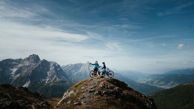 Mountain Bike Holidays © Desire Lines - Tobias Dörr Zwei Mountainbiker auf einem Berggipfel mit Ausblick auf Gebirge