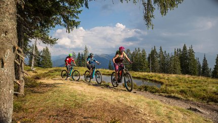 Johannes Waterfall Tour © Heiko Mandl Three mountain bikers riding on a mountain trail through green landscape.