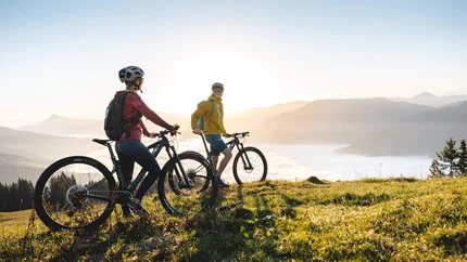BIKE TRAIL TIROL STAGE KITZBÜHEL - KIRCHBERG © TVB Kitzbüheler Alpen Brixental - Mathäus Gartner Two cyclists on a mountain overlooking sunrise and foggy valley