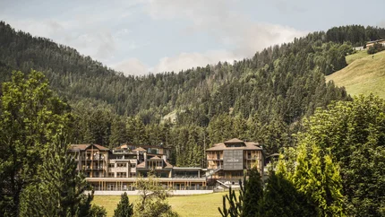 Excelsior Dolomites Life Resort © Hannes Niederkofler Hotelgebäude umgeben von Wäldern und Bergen unter bewölktem Himmel