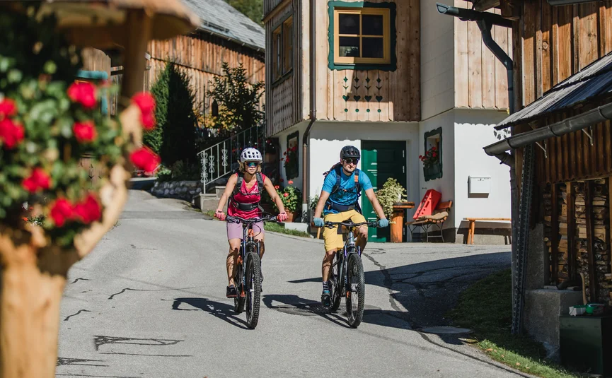 Mountain bike holidays in Styria © TVB Ausseerland Salzkammergut - Katrin Kerschbaumer Two cyclists riding through a village with wooden houses in sunny weather