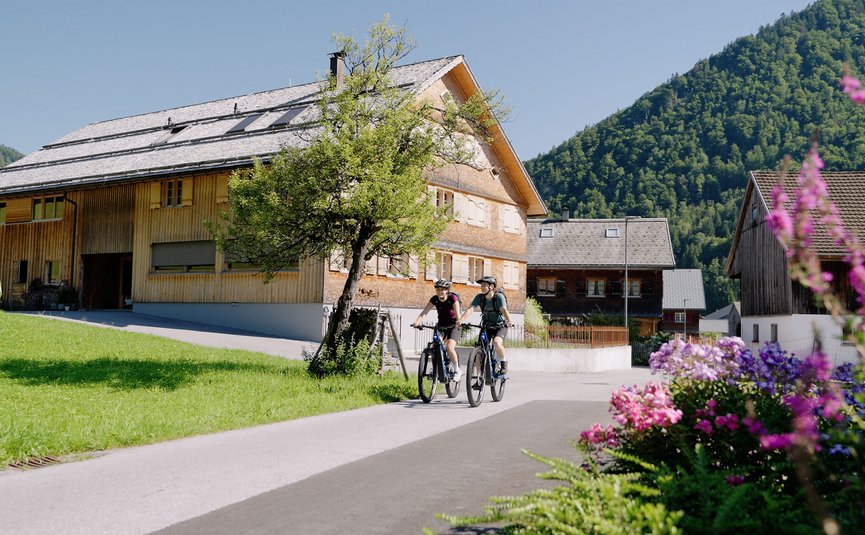 Mountain bike holiday in the Bregenzerwald © Niklas Kirchler Two cyclists riding through a village with mountains on a sunny day
