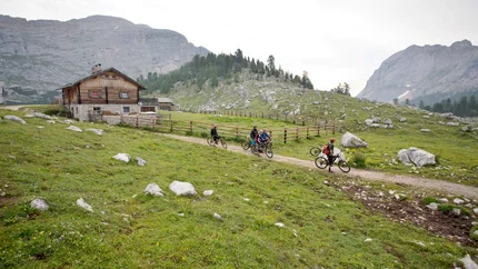ARMENTARA MEADOWS © Excelsior Dolomites Life Resort Cyclists on a path near a mountain cabin in the Alps
