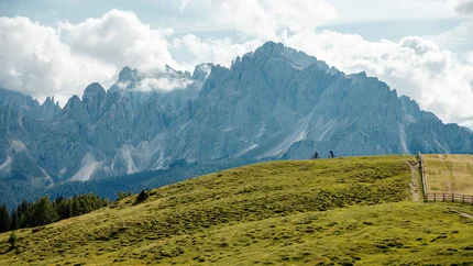 Demuth Passage © David Karg Two cyclists on green hill with towering mountains and cloudy sky