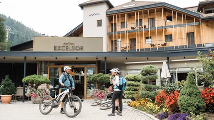 Excelsior Dolomites Life Resort © Design Innovation Award DI-A Zwei Radfahrer mit Helmen vor dem Hotel Excelsior mit blühendem Garten