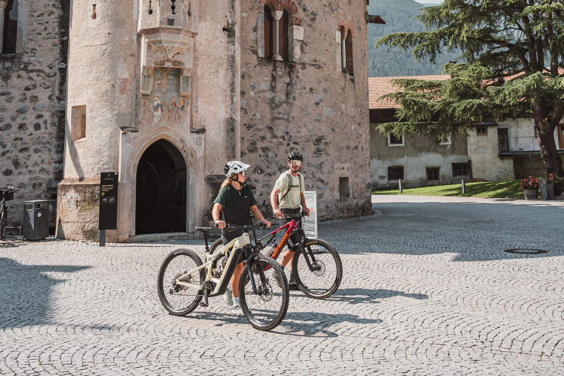 Mountain Bike Holidays © Brixen Tourismus - Hannes Engl Zwei Radfahrer vor einem historischen steinernen Turm an einem sonnigen Tag
