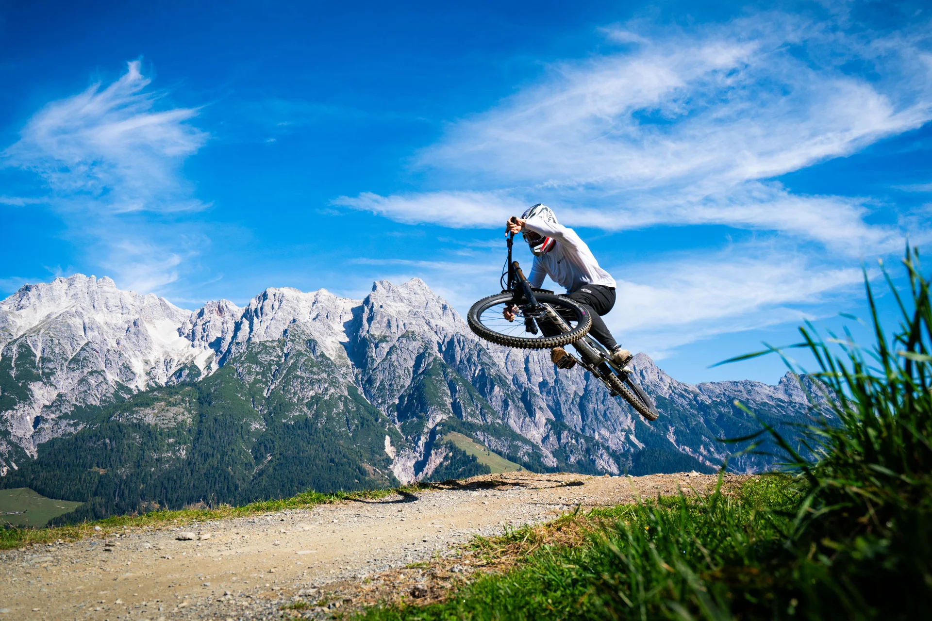 Mountain Bike Holidays © Elements Outdoor Sports Mountainbiker macht einen Sprung auf einem Bergweg mit Alpen im Hintergrund