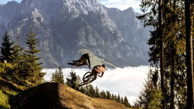 Bikepark Leogang © Klemens König Zwei Mountainbiker springen im Wald vor Bergkulisse über eine Rampe