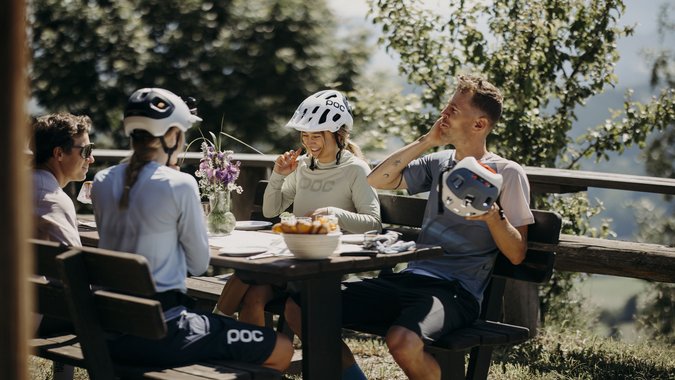 Mountain Bike Holidays © Matthias Warter Gruppe von Radfahrern mit Helmen sitzt draußen an einem Holztisch und unterhält sich