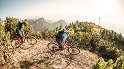 KING STAGE TO THE ROEN © WOM Medien Two mountain bikers riding on a mountain trail with mountains in the background.