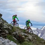 Mountain Bike Holidays © Markus Greber Zwei Mountainbiker fahren an einem steilen Berg mit schneebedeckten Gipfeln im Hintergrund